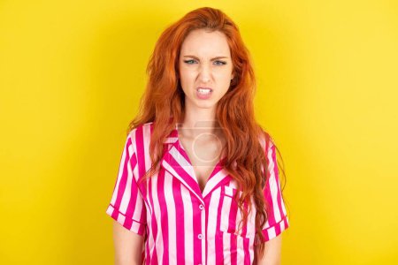 Photo for Red haired woman wearing pink pyjama over yellow studio background keeps teeth clenched, frowns face in dissatisfaction, irritated because of much duties. - Royalty Free Image