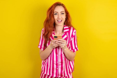 Photo for Excited red haired woman wearing pink pyjama over yellow studio background holding smartphone and looking amazed to the camera after receiving good news. - Royalty Free Image