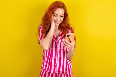 Téléchargez les photos : Femme aux cheveux rouges portant pyjama rose sur fond jaune studio tenant dans les mains sms de lecture cellulaire en utilisant la nouvelle application 5g - en image libre de droit