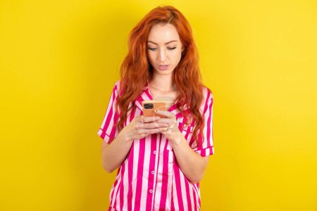 Téléchargez les photos : Focalisée femme aux cheveux rouges portant un pyjama rose sur fond jaune studio utiliser smartphone lire les nouvelles des médias sociaux, ou e-mail important - en image libre de droit