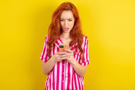 Photo for Surprised red haired woman wearing pink pyjama over yellow studio background using smartphone reading social media news, or important e-mail - Royalty Free Image