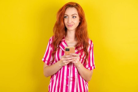 Photo for Red haired woman wearing pink pyjama over yellow studio background holds telephone hands reads good youth news look empty space advert - Royalty Free Image