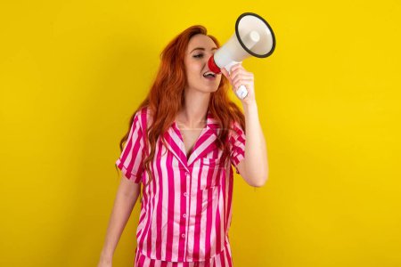 Photo for Red haired woman wearing pink pyjama over yellow studio background talking through Megaphone with Available Copy Space - Royalty Free Image