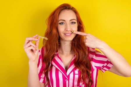 Photo for Red haired woman wearing pink pyjama over yellow studio background holding an invisible aligner and pointing to her perfect straight teeth. Dental healthcare and confidence concept. - Royalty Free Image