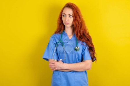 Photo for Charming thoughtful young red-haired doctor woman stands with arms folded concentrated somewhere with pensive expression thinks what to do - Royalty Free Image
