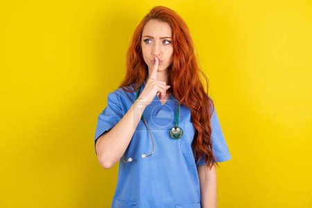 Téléchargez les photos : Jeune rousse médecin femme silence geste garde index aux lèvres fait signe de silence. Demande de ne pas partager le secret - en image libre de droit