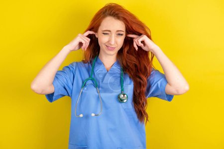 Téléchargez les photos : Heureuse jeune femme médecin aux cheveux roux ignore la musique forte et bouche les oreilles avec les doigts, demande d'éteindre le son - en image libre de droit