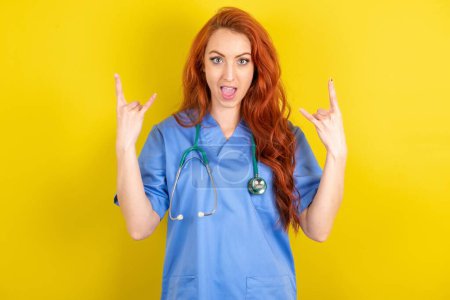 Photo for Young red-haired doctor woman makes rock n roll sign looks self-confident and cheerful enjoys cool music at party. Body language concept. - Royalty Free Image