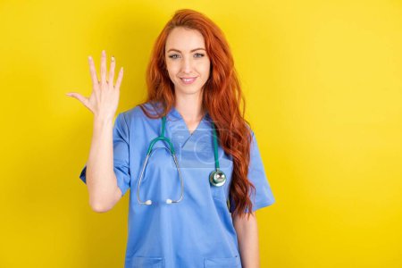 Téléchargez les photos : Jeune femme médecin rousse souriante et amicale, montrant numéro cinq ou cinquième avec la main vers l'avant, compte à rebours - en image libre de droit
