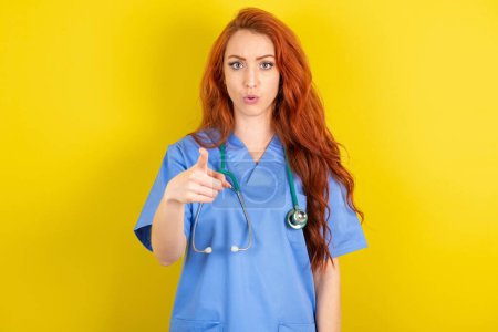 Photo for Shocked young red-haired doctor woman points at you with stunned expression - Royalty Free Image