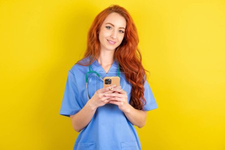 Téléchargez les photos : Jeune femme médecin aux cheveux roux Utilisation de téléphone mobile, dactylographier sms message - en image libre de droit