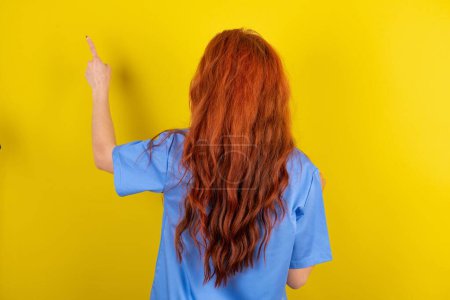 Téléchargez les photos : Jeune femme médecin aux cheveux roux pointant pour objecter sur l'espace de copie, vue arrière. Tourne le dos - en image libre de droit