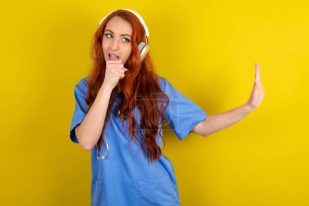 Photo for Happy young red-haired doctor woman sings favorite song keeps hand near mouth as if microphone wears wireless headphones, listens music - Royalty Free Image