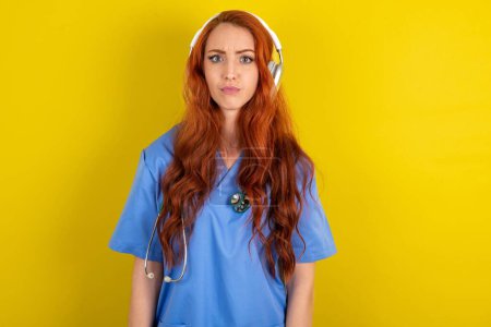 Photo for Serious displeased young red-haired doctor woman  looks puzzled at camera being angry wears stereo headphones listens music - Royalty Free Image