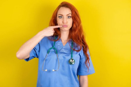 Photo for Charming young red-haired doctor woman  pointing on pout lips with forefinger, showing effect after lifting procedure, - Royalty Free Image