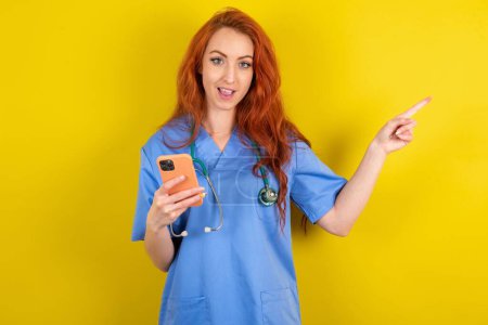 Téléchargez les photos : Étonné jeune femme médecin aux cheveux roux tenant son téléphone et pointant du doigt à côté de l'espace de copie vide - en image libre de droit