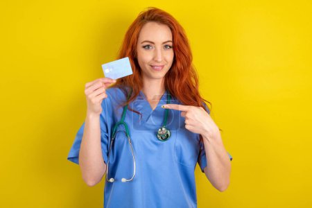 Téléchargez les photos : Portrait photo d'une jeune femme médecin rousse effectuant un achat avec une carte bancaire de crédit pointant du doigt - en image libre de droit