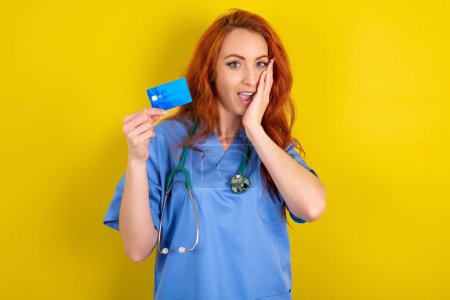 Téléchargez les photos : Étonné positif jeune femme médecin aux cheveux roux avec bras main tenir la banque de cartes en plastique fonctions impressionnées - en image libre de droit