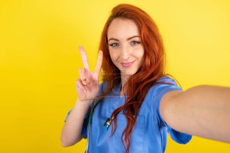 Téléchargez les photos : Positif jeune femme rousse médecin prendre selfie faire v-signe - en image libre de droit
