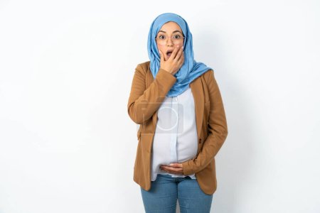 Téléchargez les photos : Femme enceinte musulmane émotionnelle portant hijab halètements de l'étonnement, couvre la bouche ouverte avec la paume, semble choqué à la caméra. - en image libre de droit