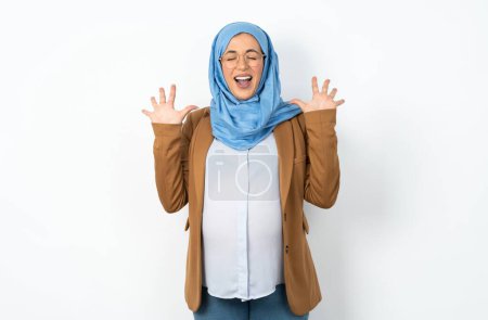 Téléchargez les photos : Femme enceinte musulmane émotive portant hijab rit bruyamment, entend blague drôle ou histoire, soulève les paumes avec satisfaction, - en image libre de droit