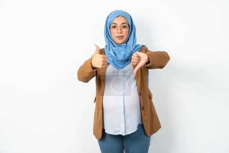 Foto de Musulmana embarazada usando hijab mostrando pulgares hacia arriba y pulgares hacia abajo, difícil elegir concepto - Imagen libre de derechos