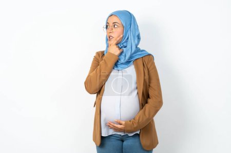 Foto de Musulmana embarazada vistiendo hijab pensando preocupado por una pregunta, preocupado y nervioso con la mano en la barbilla. - Imagen libre de derechos