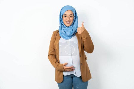Foto de Mujer musulmana embarazada usando hiyab haciendo un gesto feliz pulgares hacia arriba con la mano. Aprobación de la expresión mirando a la cámara mostrando éxito. - Imagen libre de derechos