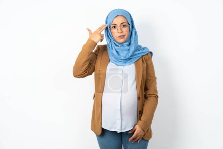 Photo for Unhappy muslim pregnant woman wearing hijab makes suicide gesture and imitates gun with hand, curves lips, keeps two fingers on temple - Royalty Free Image