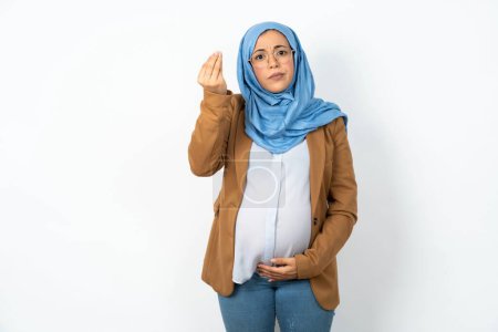 Photo for Unhappy muslim pregnant woman wearing hijab makes suicide gesture and imitates gun with hand, curves lips keeps two fingers on temple, shoots, being tired of everything, - Royalty Free Image