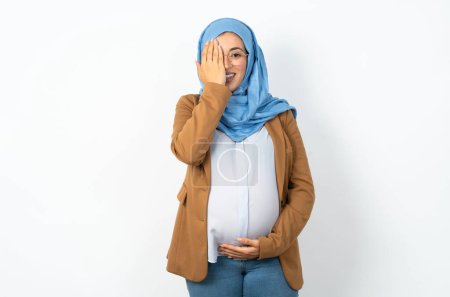 Téléchargez les photos : Musulmane enceinte femme portant hijab couvrant un oeil avec sa main, sourire confiant sur le visage et l'émotion surprise. - en image libre de droit