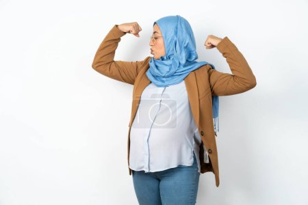 Téléchargez les photos : Femme enceinte musulmane portant hijab montrant les muscles des bras souriant fier. Concept de fitness. - en image libre de droit