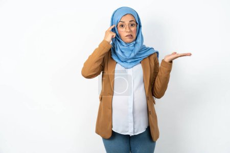 Téléchargez les photos : Femme enceinte musulmane portant hijab confus et agacé avec la paume ouverte montrant l'espace de copie et pointant du doigt vers le front. Penses-y.. - en image libre de droit