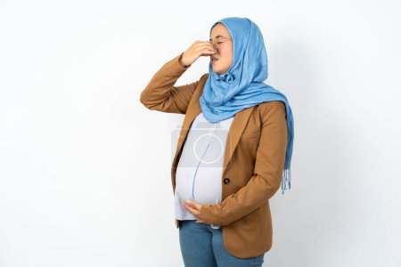 Téléchargez les photos : Femme enceinte musulmane portant hijab sentir quelque chose puant et dégoûtant, odeur intolérable, retenant souffle avec les doigts sur le nez. Mauvaise odeur - en image libre de droit