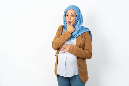 Foto de Musulmana embarazada vistiendo hijab buscando fascinado con la incredulidad, sorpresa y expresión asombrada con las manos en la barbilla - Imagen libre de derechos