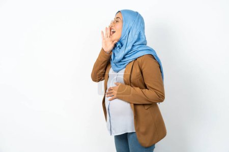 Foto de Mujer musulmana embarazada usando hijab gritando y gritando fuerte a un lado con la mano en la boca. Concepto de comunicación. - Imagen libre de derechos