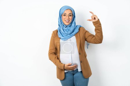 Téléchargez les photos : Femme enceinte musulmane portant hijab geste souriant et confiant avec la main faisant signe de petite taille avec les doigts à la recherche et la caméra. Concept de mesure - en image libre de droit