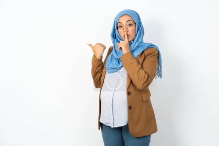 Téléchargez les photos : Femme musulmane enceinte portant hijab demandant à être calme avec le doigt sur les lèvres pointant avec la main sur le côté. Silence et concept secret. - en image libre de droit