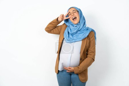 Téléchargez les photos : Femme musulmane enceinte portant hijab Faire symbole de paix avec les doigts sur le visage, souriant joyeuse montrant la victoire - en image libre de droit