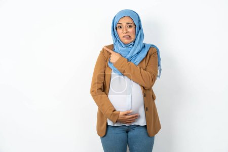 Téléchargez les photos : Femme musulmane enceinte portant hijab Pointant de côté inquiet et nerveux avec index, préoccupation et concept surprise. - en image libre de droit
