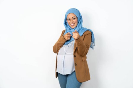 Téléchargez les photos : Belle femme musulmane enceinte portant hijab pointant les doigts vers la caméra avec un visage heureux et drôle. Bonne énergie et vibes. - en image libre de droit