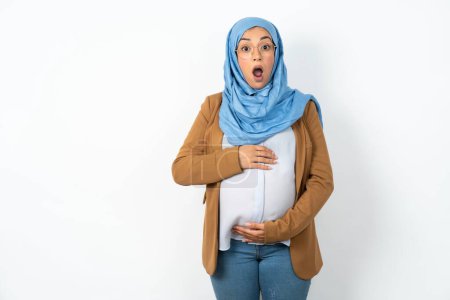 Foto de Oh, Dios mío. Sorprendido hermosa mujer musulmana embarazada con hijab mira fijamente a la cámara con expresión conmocionada exclama con inesperada, - Imagen libre de derechos
