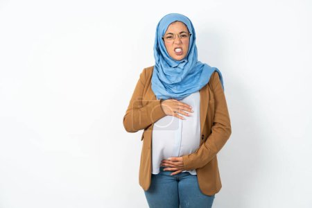 Foto de Hermosa mujer musulmana embarazada usando hijab mantiene los dientes apretados, frunce el ceño cara en la insatisfacción, irritado debido a muchos deberes. - Imagen libre de derechos