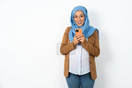 Photo for Beautiful pregnant muslim woman wearing hijab  holding a smartphone and looking sideways at blank copyspace. - Royalty Free Image