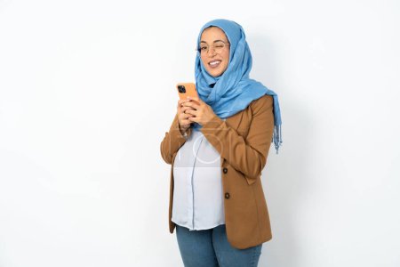 Foto de Emocionada mujer musulmana embarazada con guiños hijab y uso de teléfonos inteligentes ojo hold leer noticias de redes sociales - Imagen libre de derechos