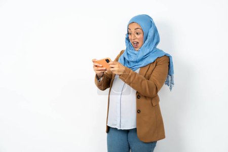 Photo for Portrait of an excited pregnant muslim woman wearing hijab  playing games on mobile phone. - Royalty Free Image