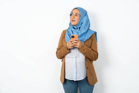 Photo for Pregnant muslim woman wearing hijab  holds telephone hands reads good youth news look empty space advert - Royalty Free Image