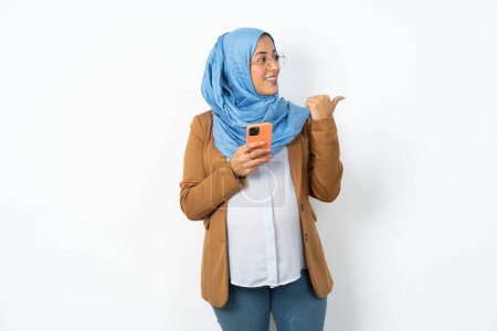 Foto de Mujer musulmana embarazada usando hijab usando y mensajes de texto con el teléfono inteligente apuntando y mostrando con el pulgar hacia el lado con la cara feliz sonriendo - Imagen libre de derechos