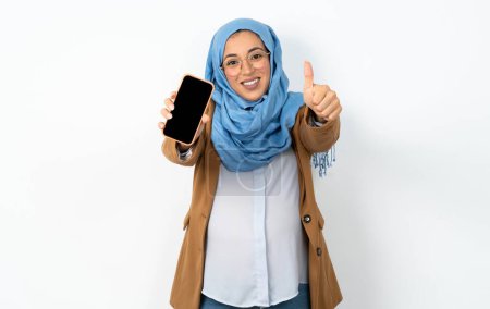 Téléchargez les photos : Femme musulmane enceinte portant hijab tenant dans la cellule de la main montrant donnant pouce écran noir vers le haut - en image libre de droit