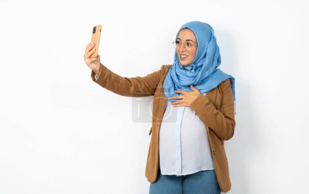 Téléchargez les photos : Femme musulmane enceinte portant le hijab souriant et prenant un selfie prêt à le poster sur ses médias sociaux. - en image libre de droit